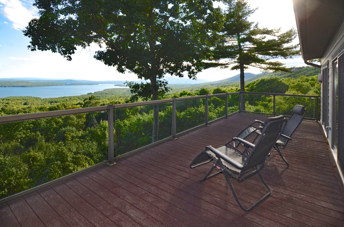 A spacious outdoor deck is furnished with reclining chairs, offering expansive views of the Ashokan Reservoir and Catskill Mountains. Lush greenery surrounds the deck, creating a serene atmosphere. The deck's railing provides safety while enhancing the feeling of openness to the surrounding landscape.