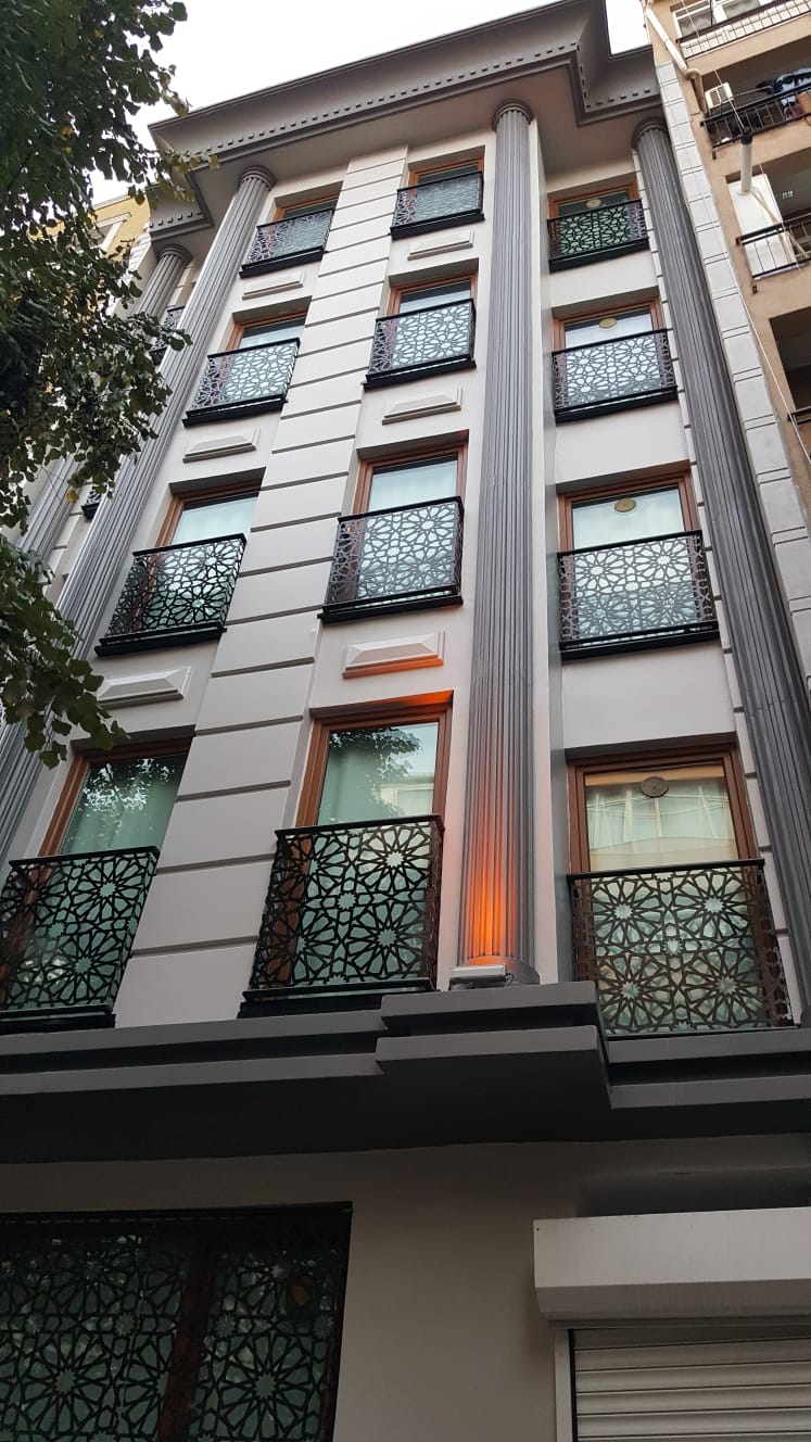 The exterior of a contemporary building is showcased, featuring intricate balcony railings and large windows. The facade displays a sleek design with vertical lines and a subtle color contrast. Soft lighting is visible by the entrance, enhancing the building's modern aesthetic.