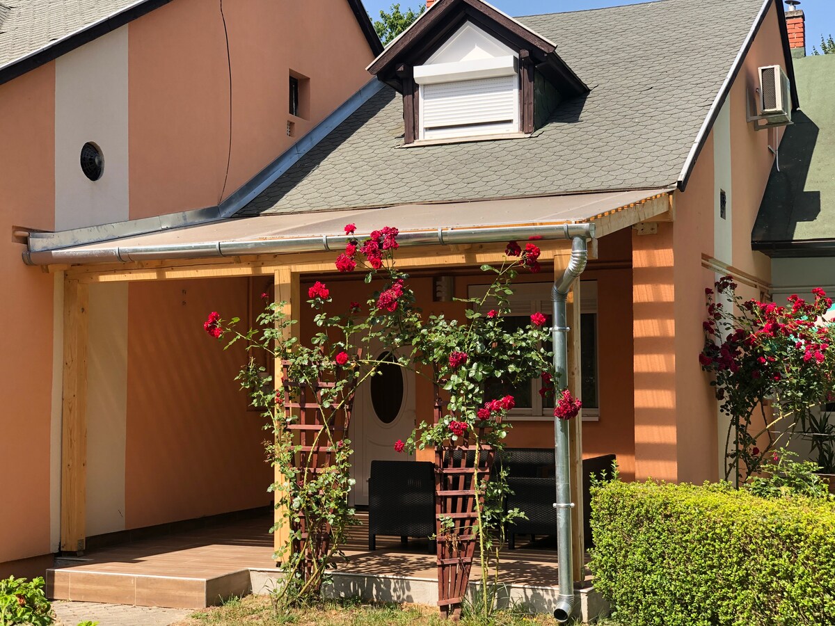 The exterior of a charming apartment is shown, featuring a covered porch adorned with climbing roses. A seating area is visible under the eaves, offering a space for relaxation. The walls are painted in warm tones, complementing the lush greenery surrounding the property.