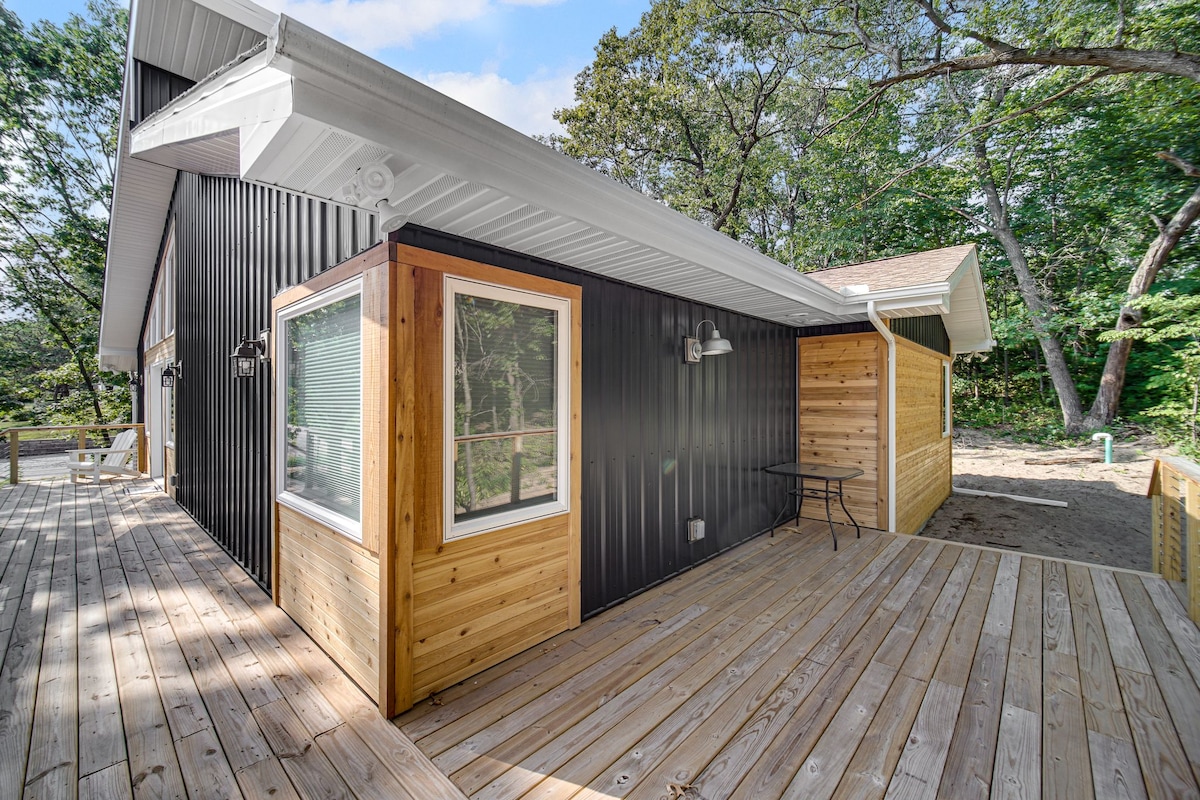 The exterior of the modern home displays a combination of sleek black and natural wood paneling. A spacious wrap-around deck is visible, accompanied by a small table and chairs set against a backdrop of trees, providing a serene and relaxing environment.