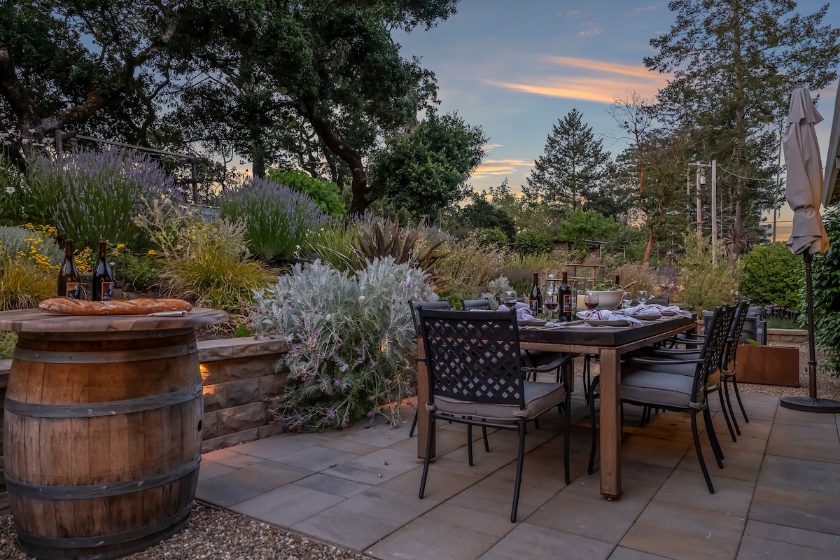 An outdoor dining area is set under the open sky, featuring a long wooden table surrounded by dark chairs. Stone planters filled with greenery and colorful flowers are present in the background, while a wine barrel and additional bottles enhance the inviting atmosphere.