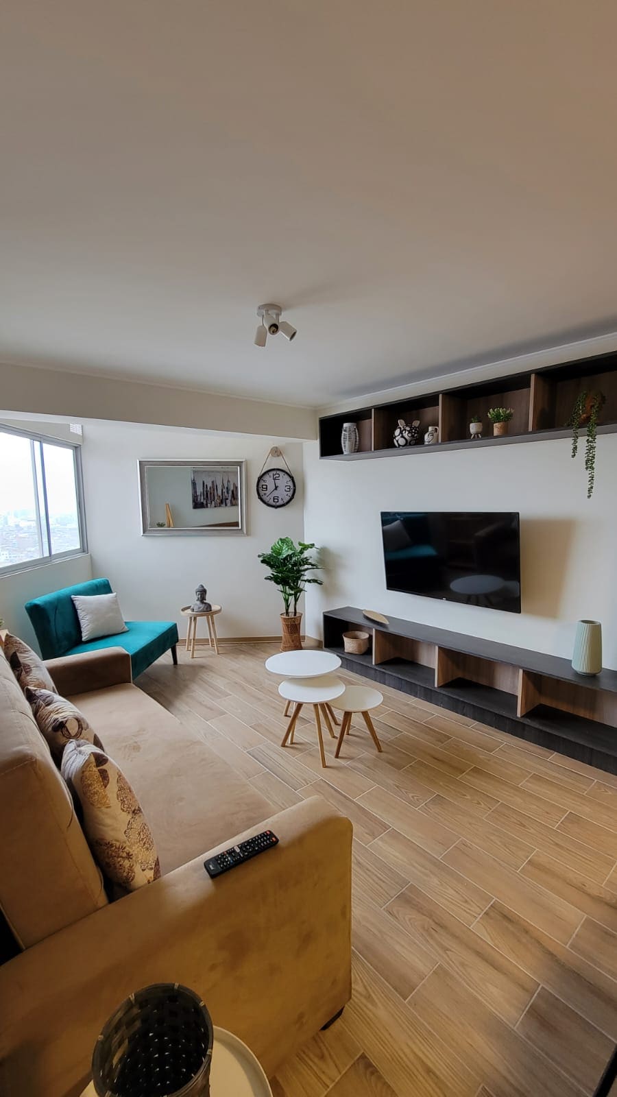 The living area features a comfortable sofa beside two round tables. A large television is mounted on the wall, accompanied by decorative shelves. Natural light enters through a window, and greenery is seen in a nearby potted plant, enhancing the space's inviting feel.