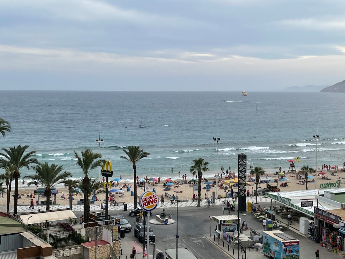 A panoramic view of the beachfront captures the sandy shore with umbrellas, sunbathers, and beach activities. Palm trees line the beach, while watercraft can be seen on the sea. In the foreground, fast-food establishments and shops are visible along the promenade.