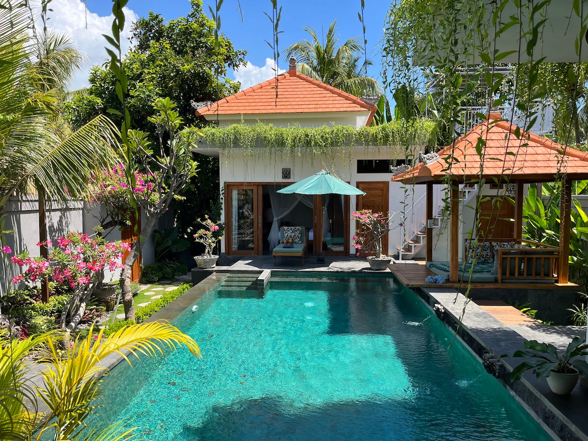 A serene outdoor space features a private pool surrounded by lush greenery. Tropical plants and flowering shrubs provide a vibrant backdrop, while a covered sitting area beside the pool invites relaxation. The villa is designed with a mix of modern and traditional Balinese architecture.