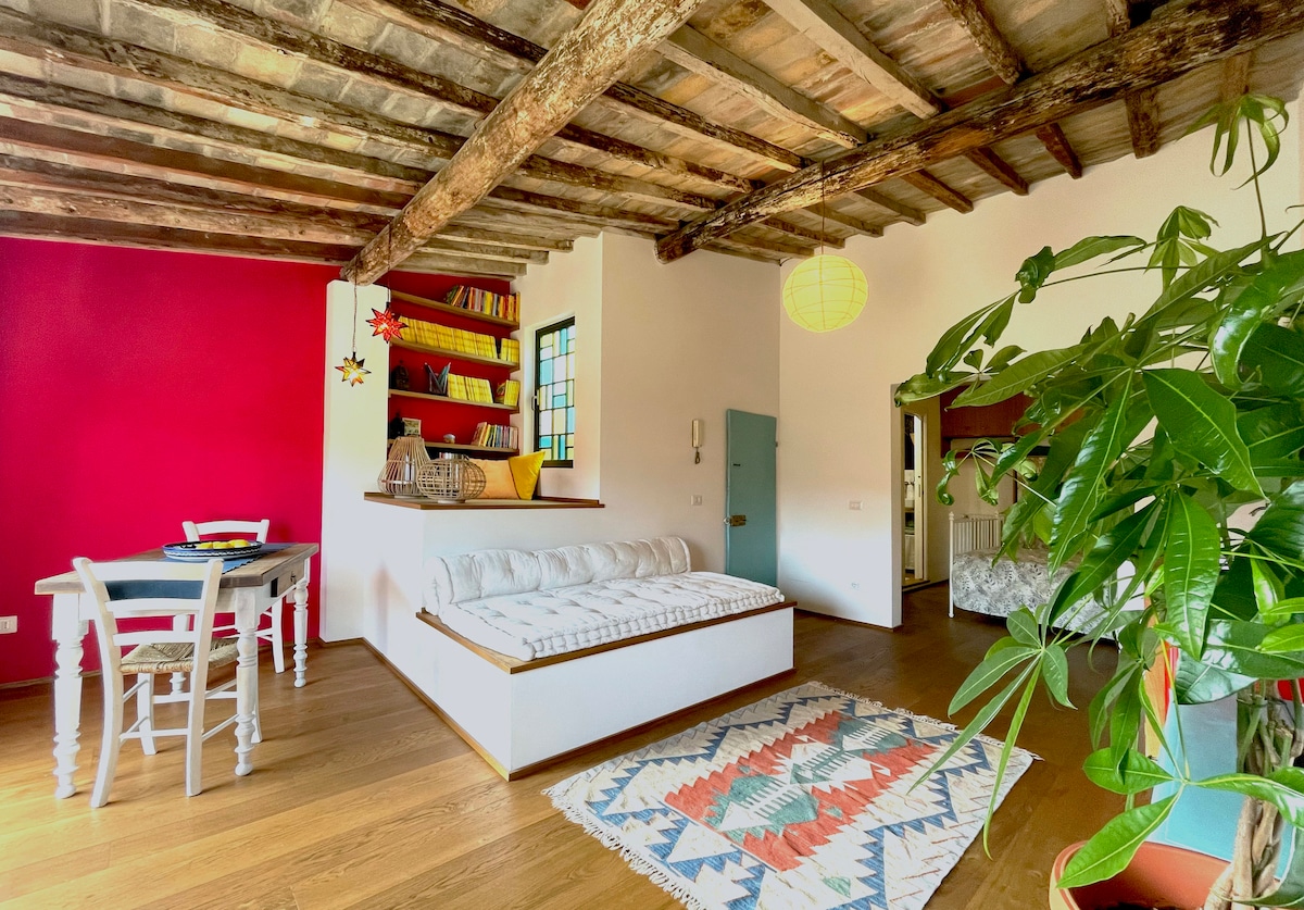 A spacious loft interior is highlighted by rustic wooden beams and striking red accent wall. A seating area with a cozy white sofa is complemented by a patterned rug. A dining table and greenery add to the inviting ambiance, while shelves display books and decor.