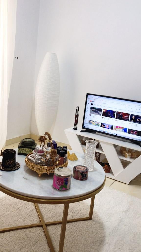 A stylish living area features a round marble coffee table adorned with various candles and a decorative tray. A modern white lamp stands beside the table, and a television is positioned on a sleek white stand, displaying an array of colorful content.