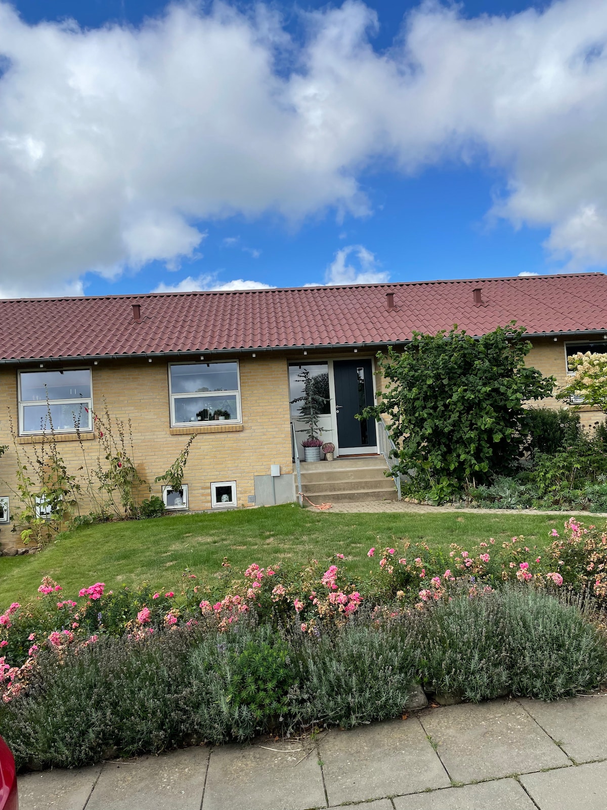 A welcoming exterior of a single-story house is shown, featuring a neatly maintained garden with colorful flowers. A stone path leads to the entrance, which is flanked by potted plants. Above, a roof with red tiles contrasts against a blue sky adorned with fluffy clouds.