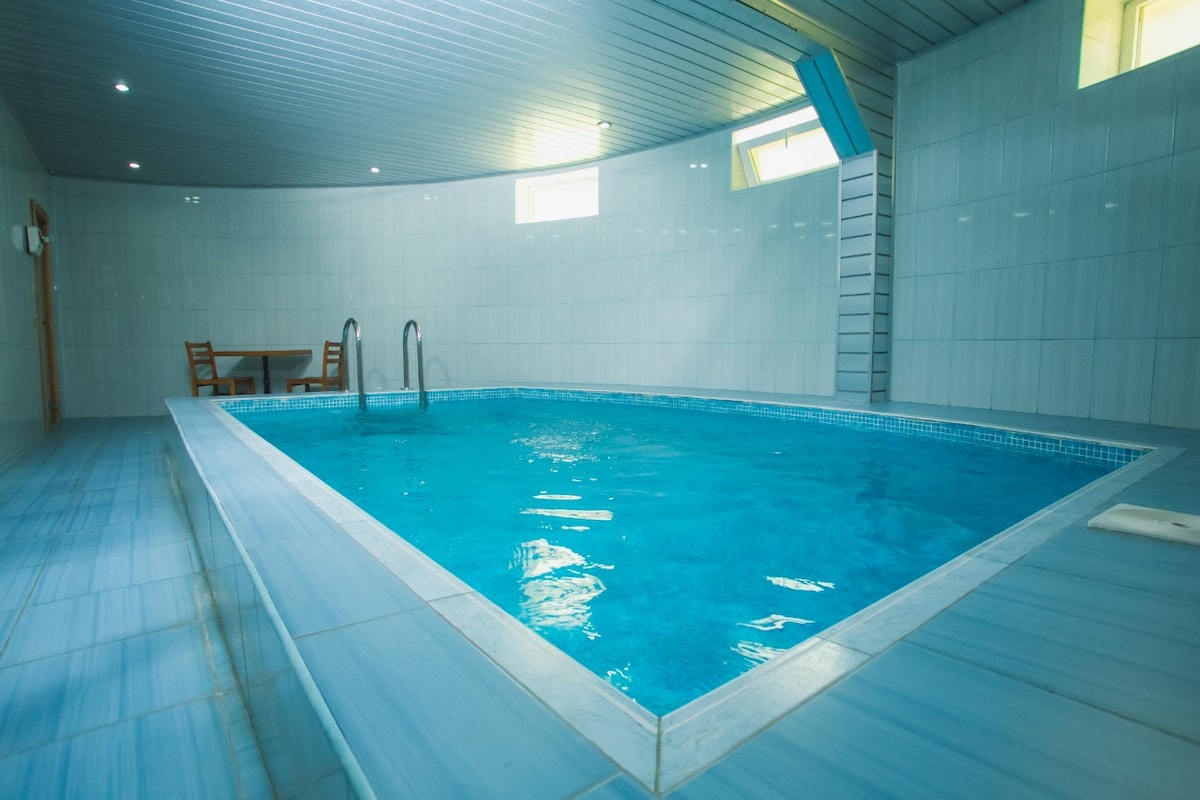 An indoor pool is showcased, featuring clear blue water and sleek tiled walls. Natural light enters through windows, creating a bright atmosphere. A wooden table and chairs are positioned nearby, providing a space for relaxation. The curved shape of the room adds a unique architectural element.