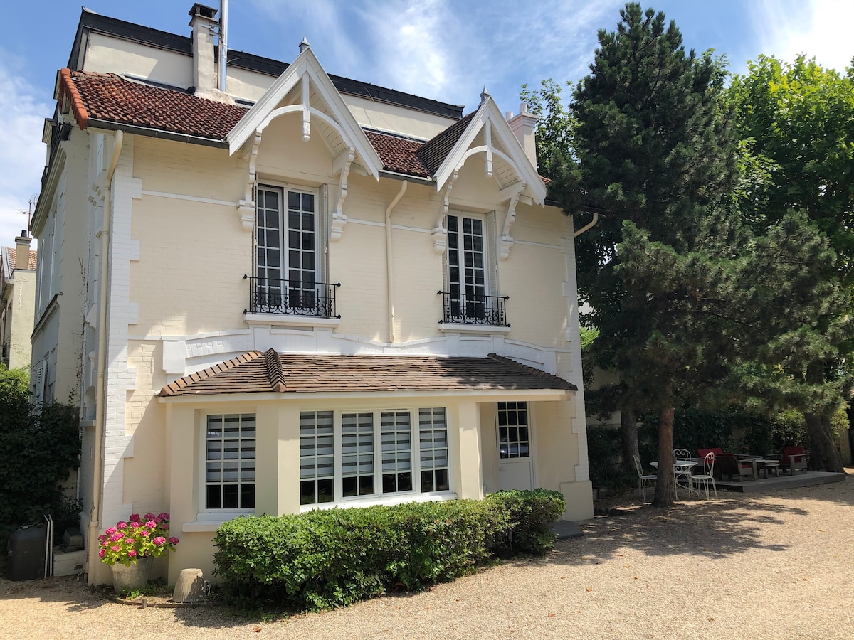 The exterior of the charming guest house features a bright façade with white and light-colored accents. Two large windows with decorative balconies create a welcoming appearance, while a shaded garden area is visible in front, lined with greenery and small flowering plants.
