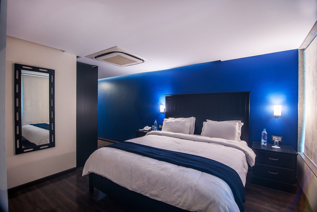 The spacious bedroom features a large king-sized bed with crisp white linens. Dark blue accent walls provide a calming backdrop. A full-length mirror is positioned adjacent to the bed. Natural light filters through the window, illuminating the room's modern decor.