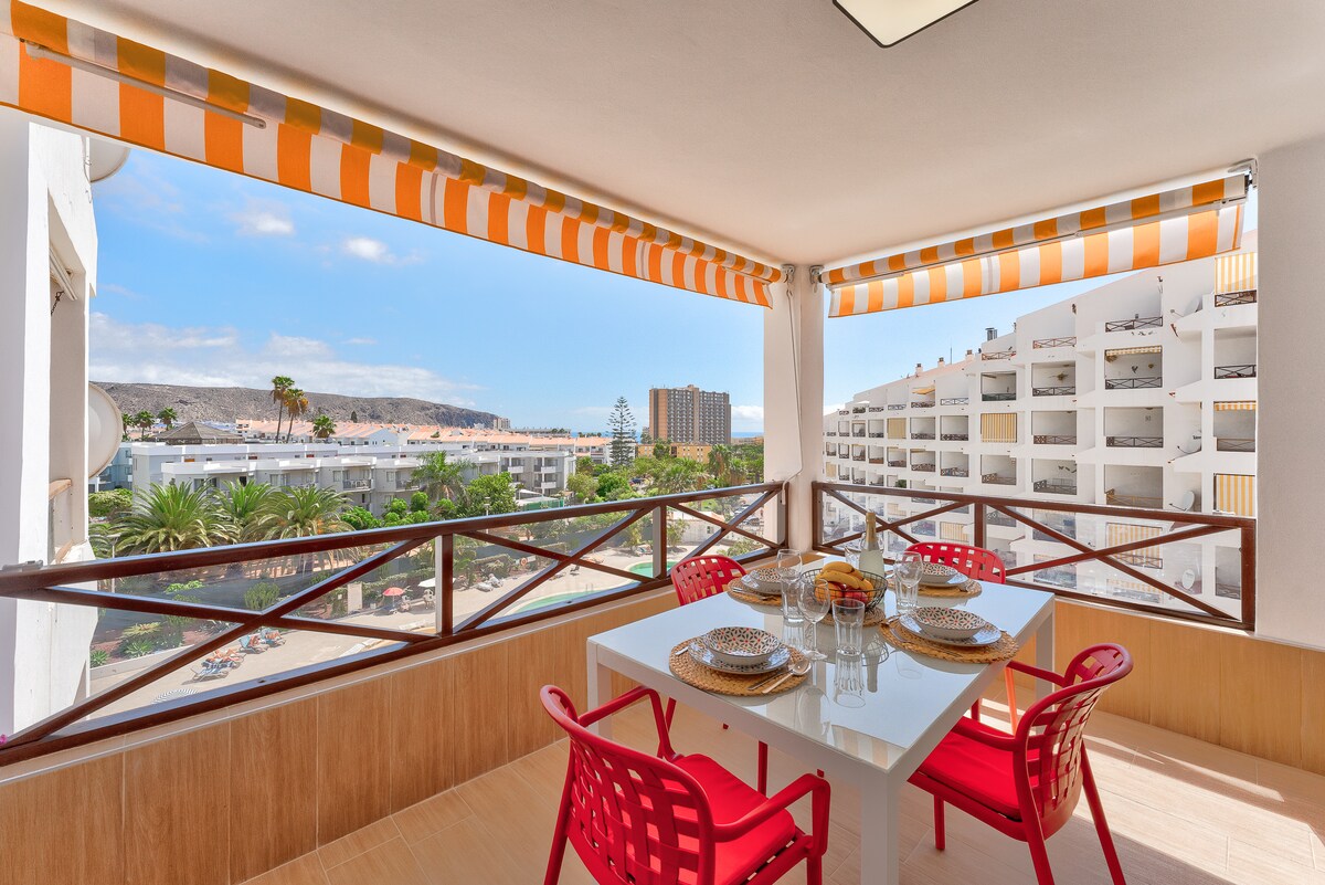 A spacious terrace is presented with a dining table and four red chairs, allowing for outdoor meals. The area is shaded by striped awnings. Scenic views showcase palm trees, neighboring buildings, and distant mountains under a clear blue sky.