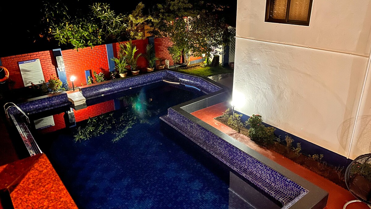 A serene outdoor pool is illuminated by soft lighting, surrounded by lush greenery and vibrant plant life. The pool features a mosaic tile design, with steps leading into the water, creating a tranquil retreat. Nearby, the walls are painted in bright colors, adding to the charm.