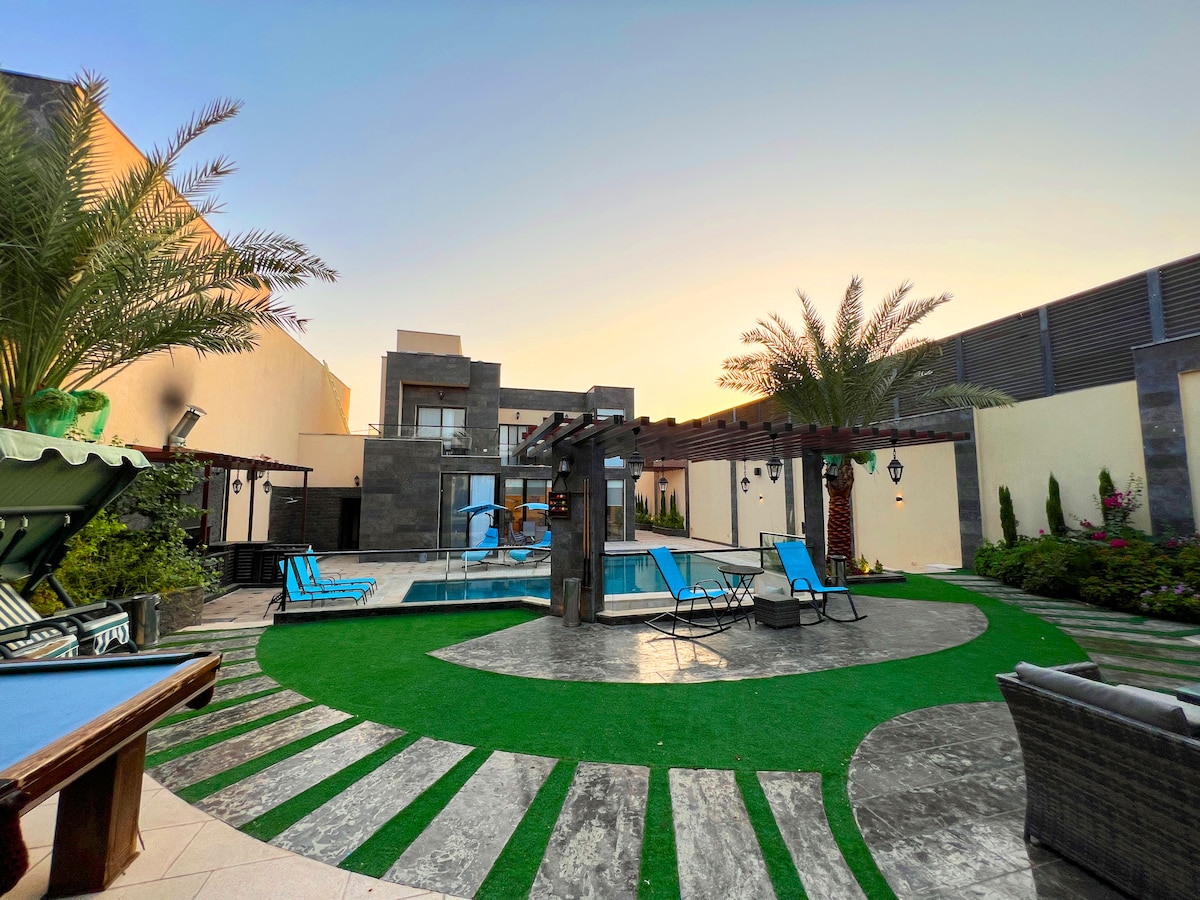 The outdoor area features a spacious garden with green grass and palm trees. Lounge chairs surround a swimming pool, creating a relaxing atmosphere. A shaded seating area is visible, complemented by a stone pathway that connects various sections of the garden.