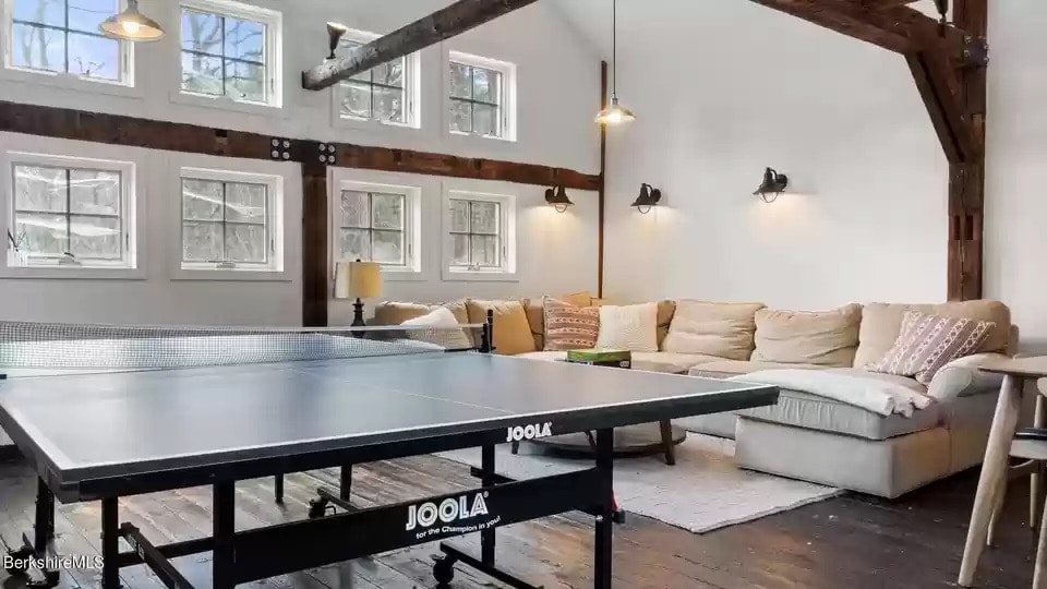 A spacious game area features a large pingpong table situated near a comfortable seating arrangement. Natural light filters through multiple windows, illuminating the room's rustic beams and cozy decor, creating an inviting space for relaxation and entertainment.