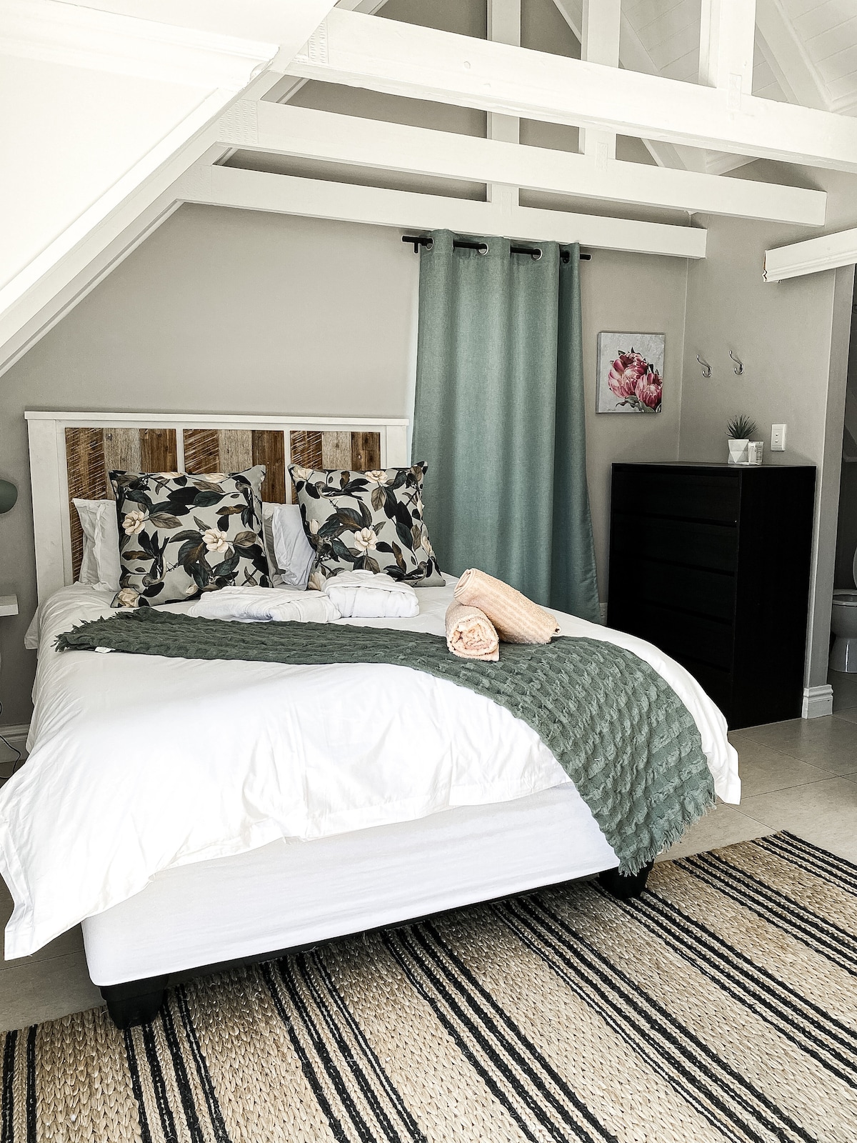 A cozy sleeping area features a spacious bed dressed in white linens, complemented by patterned throw pillows. A soft green blanket is casually arranged at the foot of the bed. A light-colored dresser stands nearby, with soft curtains framing the window.