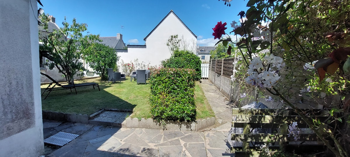 A well-maintained garden area features manicured grass bordered by flowering shrubs. Comfortable seating options are visible, along with a small stone path leading to the entrance. A clear blue sky and sunshine enhance the inviting outdoor space, ideal for relaxation.