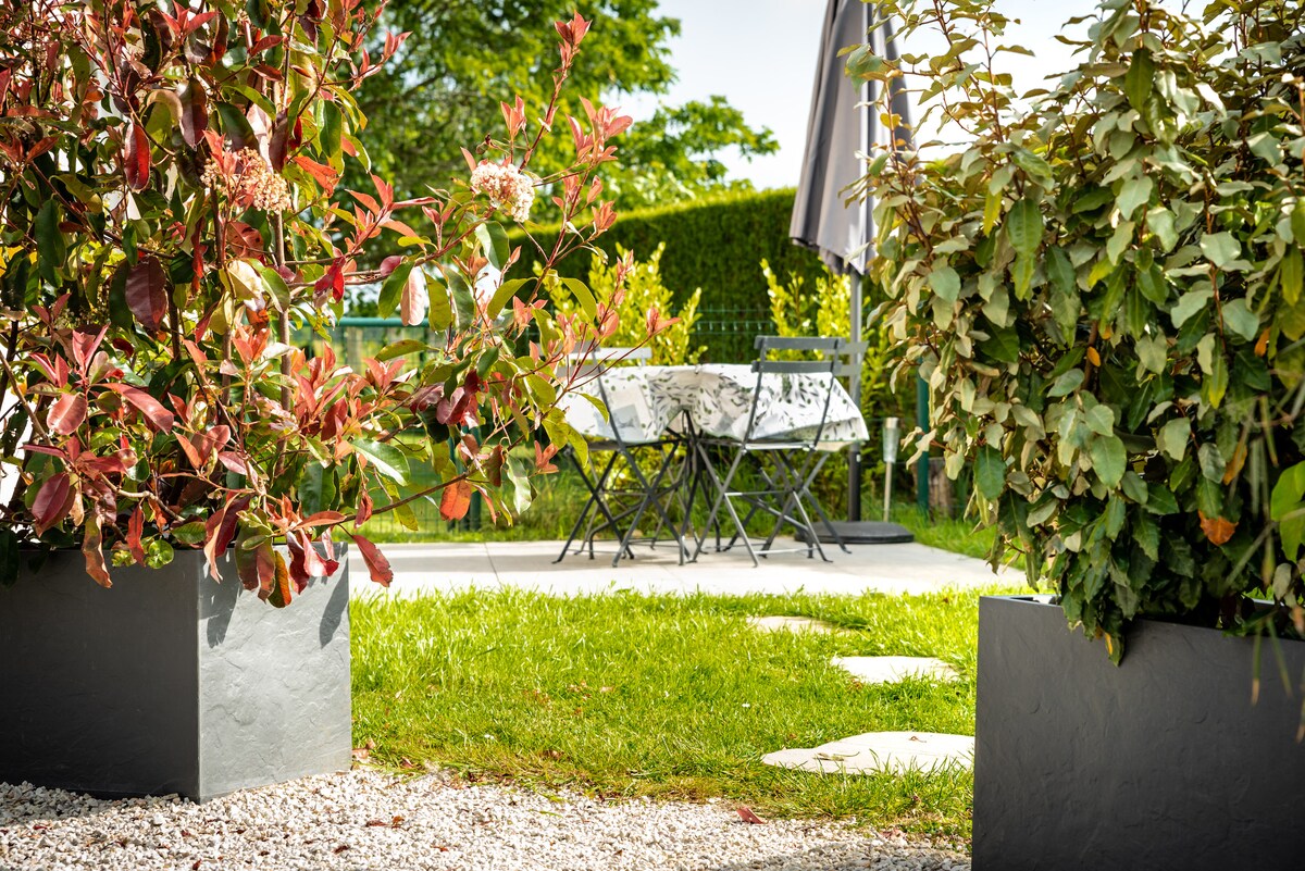 A private garden area is framed by lush greenery and vibrant foliage. A small table with two chairs is visible on a patio surface, inviting relaxation among nature. This outdoor space is designed for comfort and serenity.