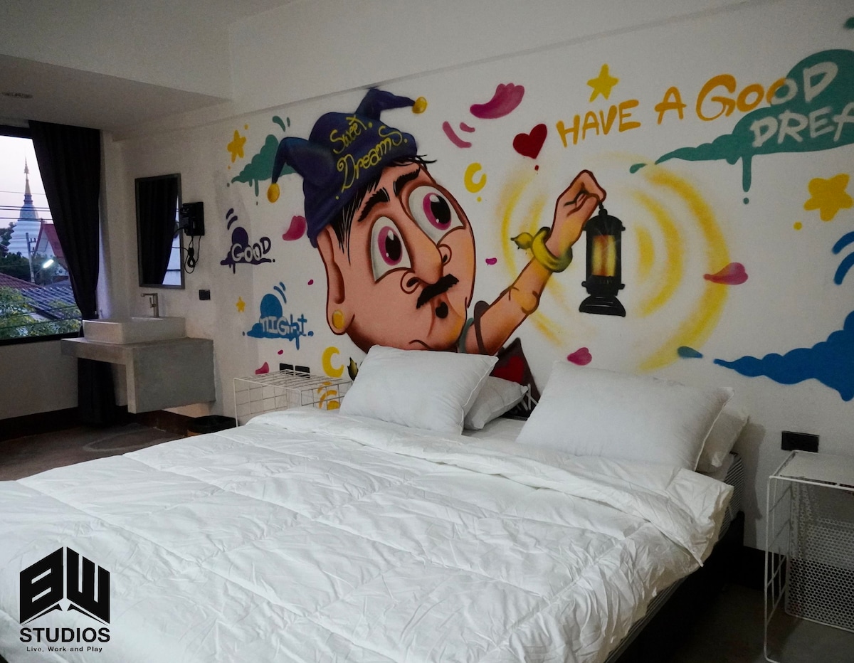 A spacious sleeping area features a large bed with crisp white linens. A vibrant mural depicting a whimsical character holding a lantern adorns one wall, adding a touch of creativity. Light can be seen streaming in from a nearby window covered with dark curtains.