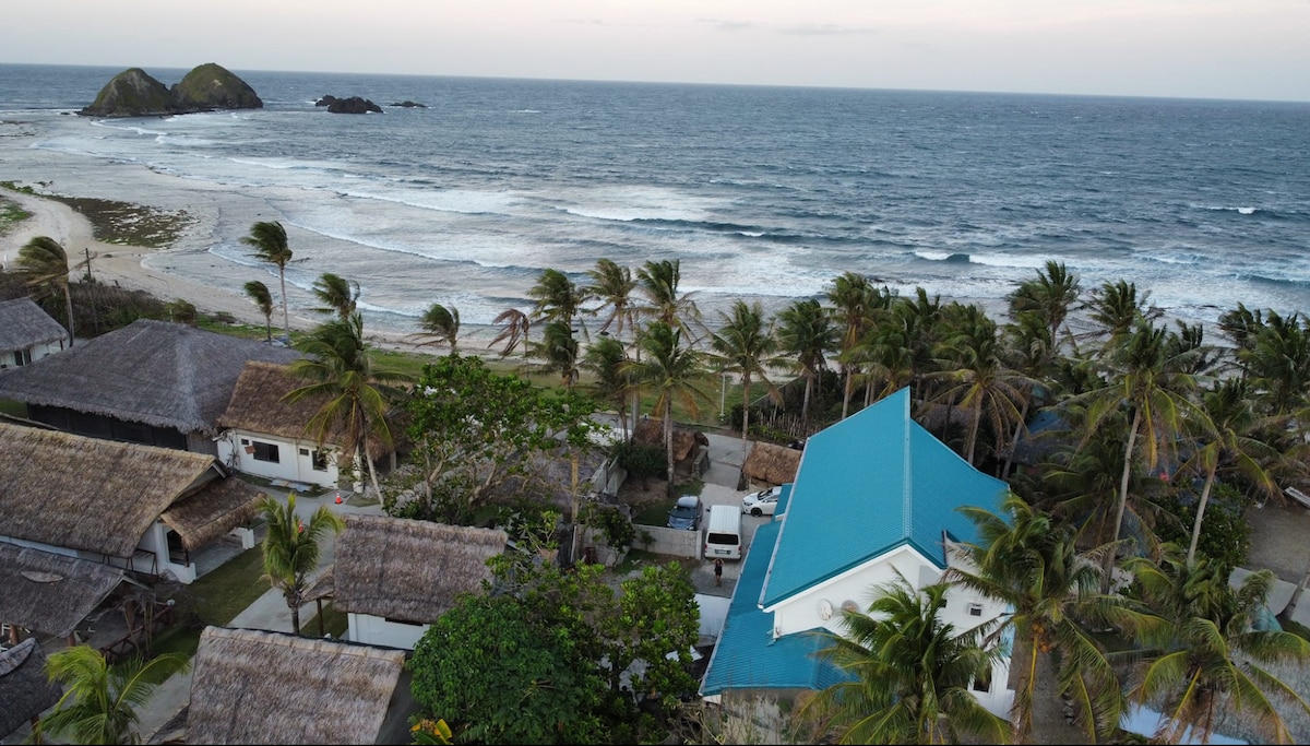 An aerial view captures a coastal landscape featuring gentle waves lapping at the shore. Palm trees sway in the breeze, framing rustic buildings with thatched roofs and a blue-roofed structure. Scenic hills rise in the distance, providing a backdrop to the serene beachfront.