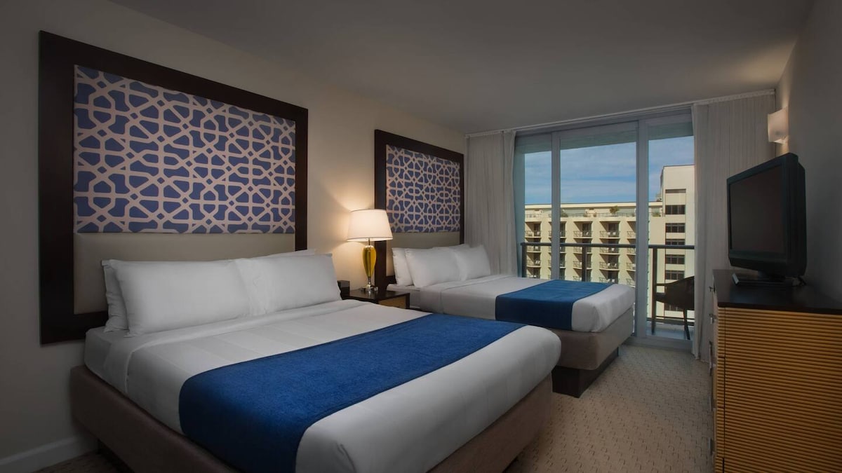 A well-appointed sleeping area features two queen beds with crisp white linens and blue accents. A vibrant art piece is displayed above each bed, and large glass doors provide natural light and access to a furnished balcony. A flat-screen television is positioned against a wooden cabinet.
