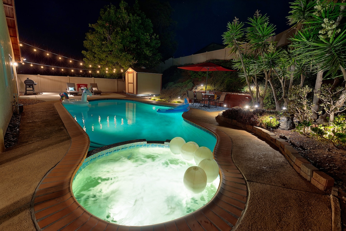 A private pool and jacuzzi area is illuminated by soft lighting, creating a calming evening ambiance. Surrounding landscaping features tropical plants and subtle lighting. A seating area with an umbrella is visible, enhancing the outdoor gathering space. Floating pool toys add a playful touch.
