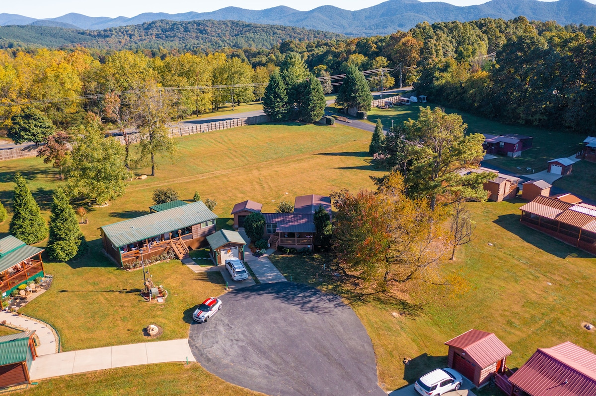 An aerial view captures the expansive landscape surrounding the cabin community. Green hills and tree-covered mountains provide a natural backdrop. The neatly arranged cabins are visible, along with well-maintained grassy areas and a paved parking space.