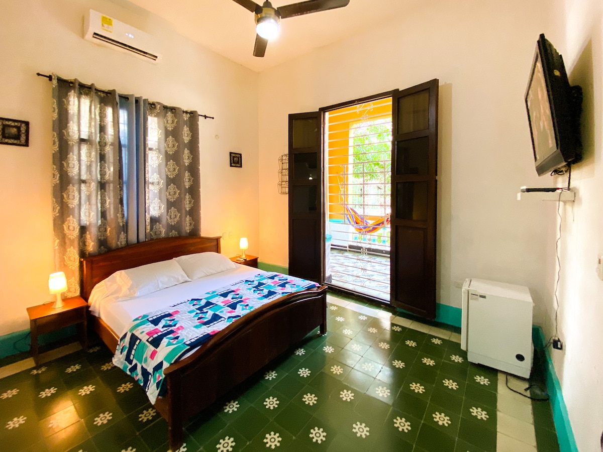 A well-lit bedroom features a spacious bed with a decorative blanket. Two bedside lamps provide soft illumination. Large windows open to a patio area, framed by vibrant curtains, allowing natural light to fill the room. A wall-mounted television is positioned above a small air conditioning unit.