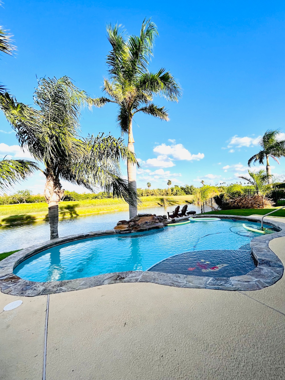 A sparkling saltwater pool is framed by lush palm trees, with a serene view of the lake in the background. Comfortable lounge chairs sit nearby on the patio, inviting relaxation under the clear blue sky.