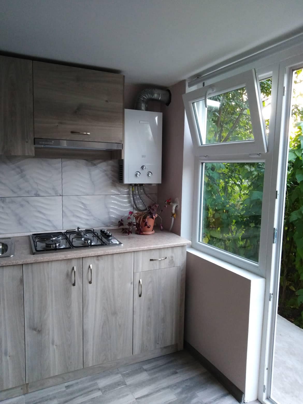A modern kitchen corner features wooden cabinetry with a blend of light and dark finishes. A gas cooktop is built into the countertop, while a bright window offers a view of greenery outside. A boiler is installed along the wall, enhancing functionality.