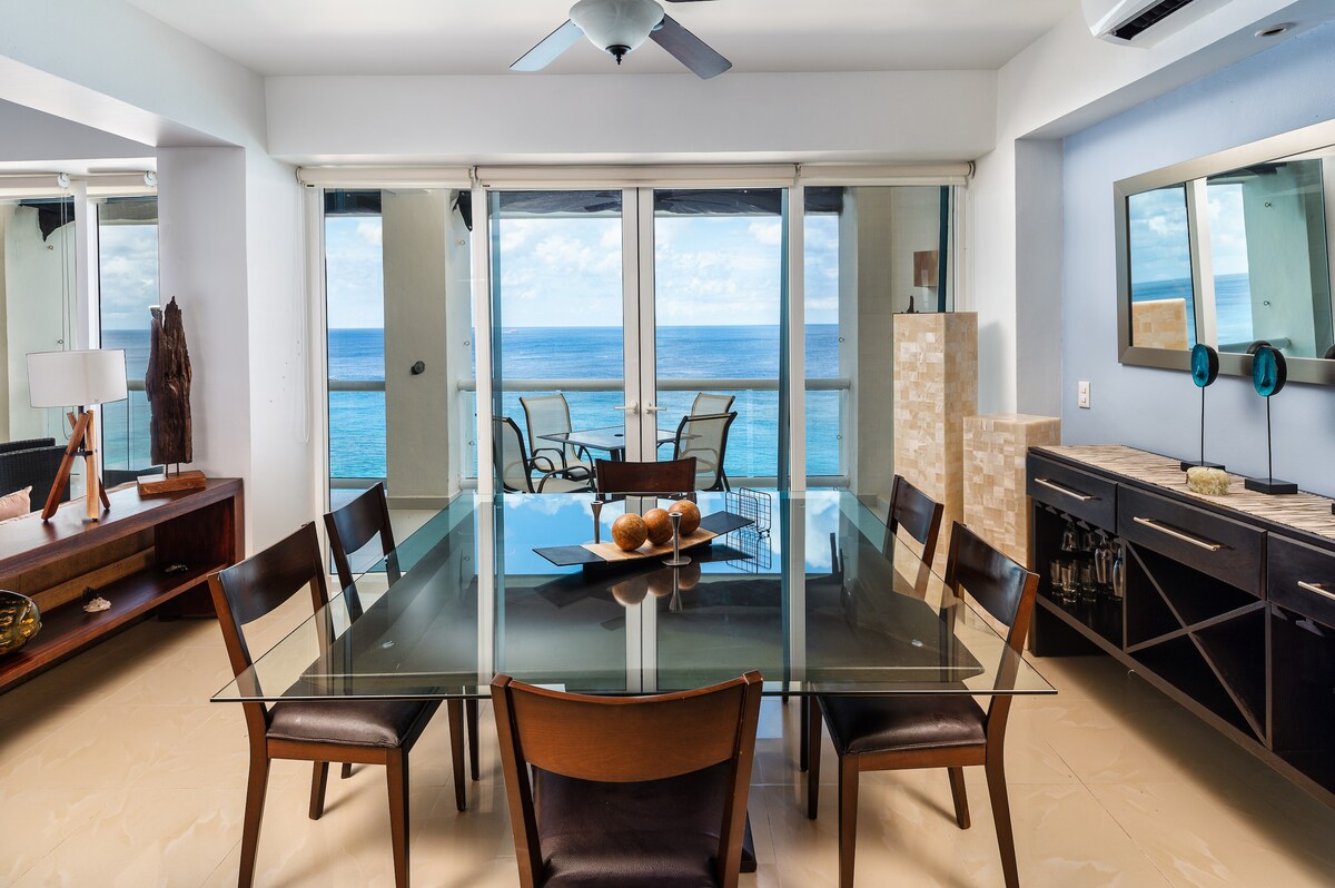The dining area features a glass-top table surrounded by wooden chairs, with a view of the ocean visible through expansive sliding glass doors. Natural light illuminates the space, highlighting the minimalist decor and the oceanfront beyond.