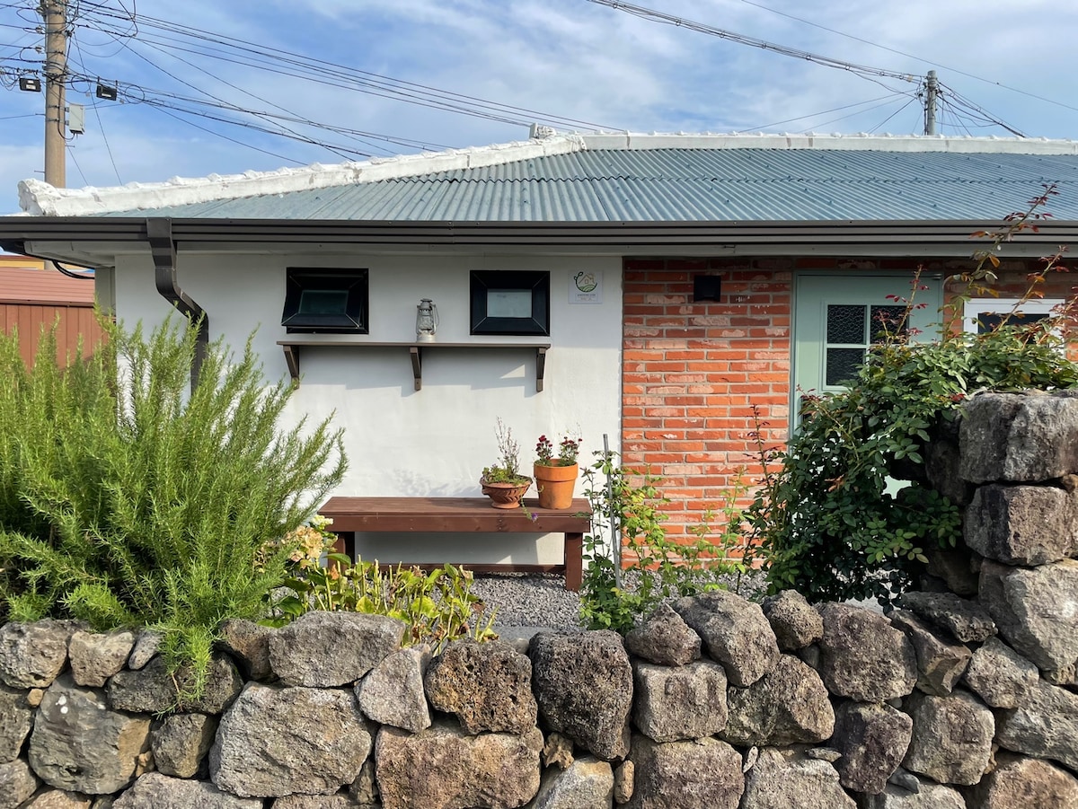 The exterior of a renovated traditional Jeju home is showcased, featuring a combination of brick and white walls. A wooden bench with potted plants is positioned in front of the house, while a rustic stone wall adds character. Green shrubs enhance the natural setting.