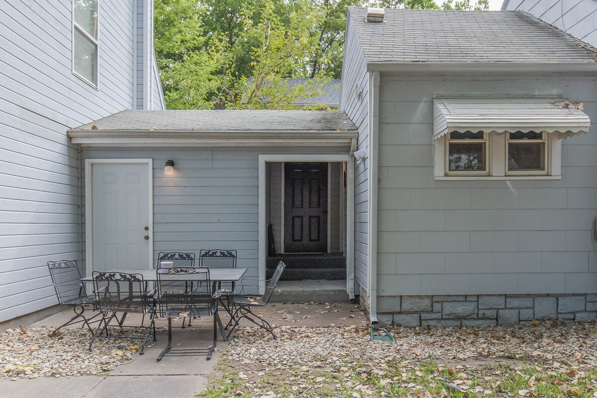 A private entrance to the basement apartment is visible, featuring a door framed by light greenery. A small patio area includes a table with four chairs, inviting outdoor dining. The entrance is marked, leading to steps that descend into the apartment.