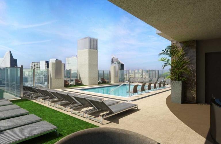 A rooftop pool area is depicted, surrounded by comfortable lounge chairs arranged neatly around the pool. Lush greenery is visible along the edge, complementing the expansive city skyline in the background under a clear blue sky.