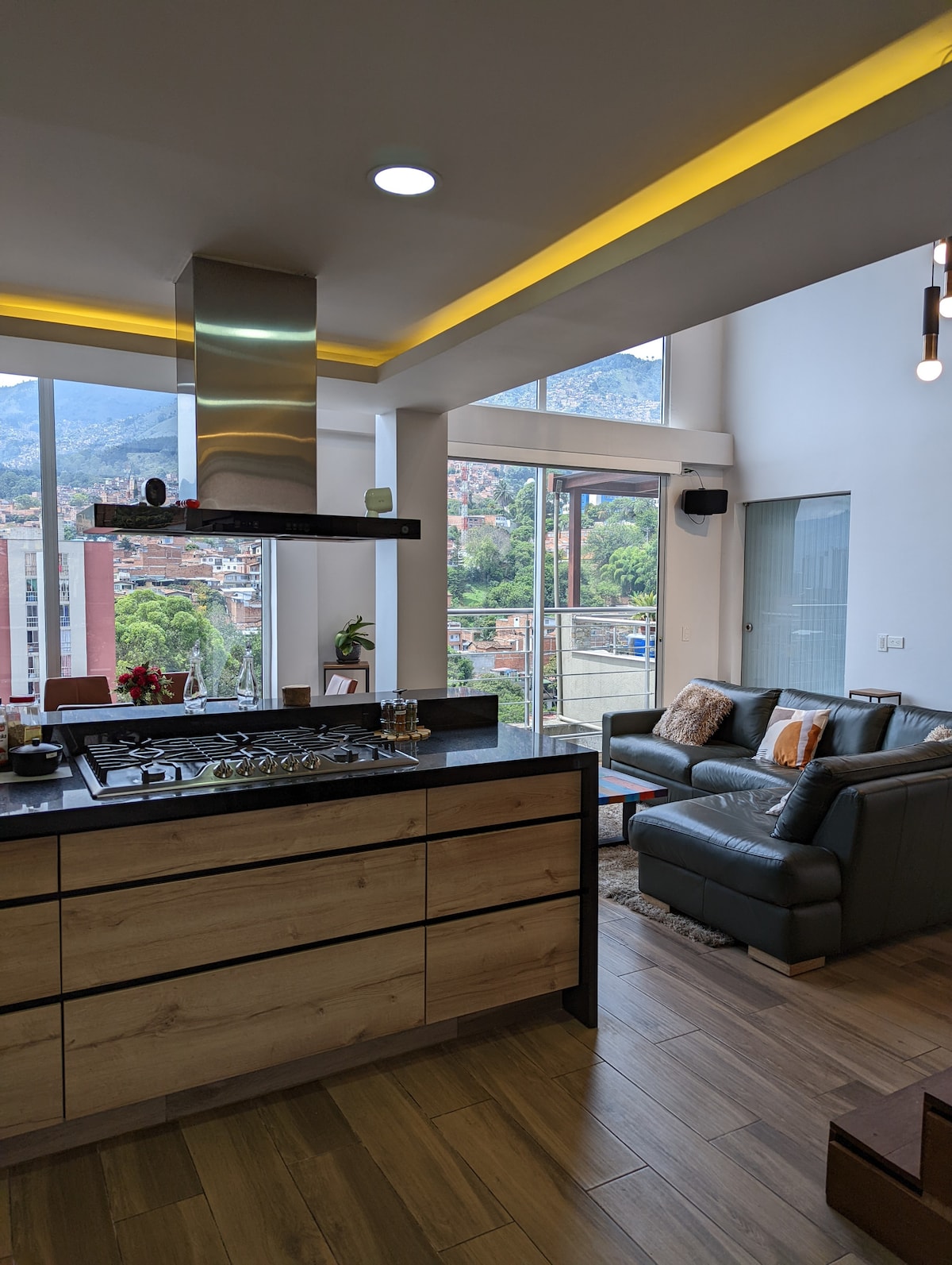 A modern kitchen area features a sleek gas stove with an overhead vent, complemented by wooden cabinetry. Natural light floods the space through large windows, revealing mountain views. A cozy seating area with a gray sofa and decorative cushions enhances the open layout.