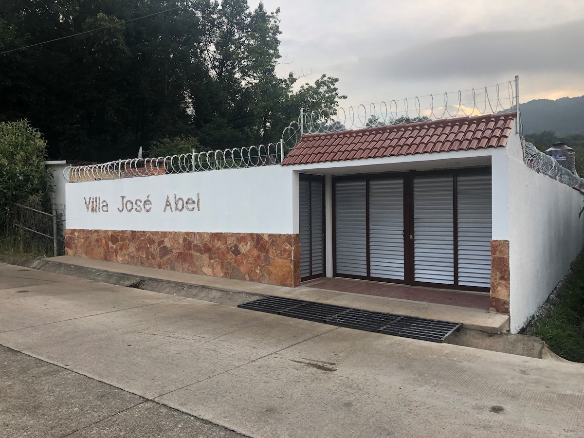 A securely fenced entrance to Villa José Abel is shown, featuring a white wall with the villa's name prominently displayed. The exterior includes a covered porch area with shuttered windows. Lush greenery from trees surrounds the property, contributing to a sense of privacy.