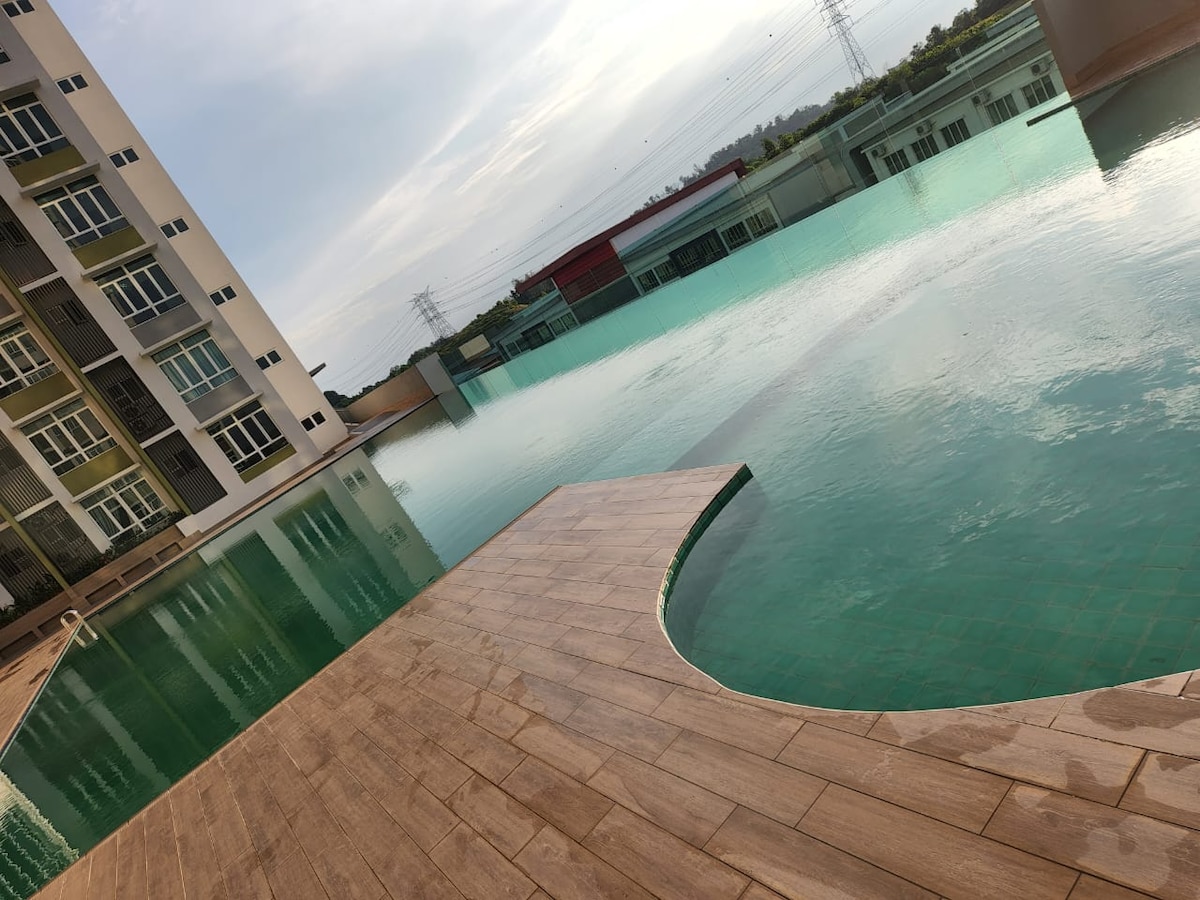 An expansive swimming pool is viewed, featuring a smooth wooden deck that curves gently. The water appears calm, reflecting the sky above. Surrounding structures are partially visible, offering a backdrop to the serene swimming area.