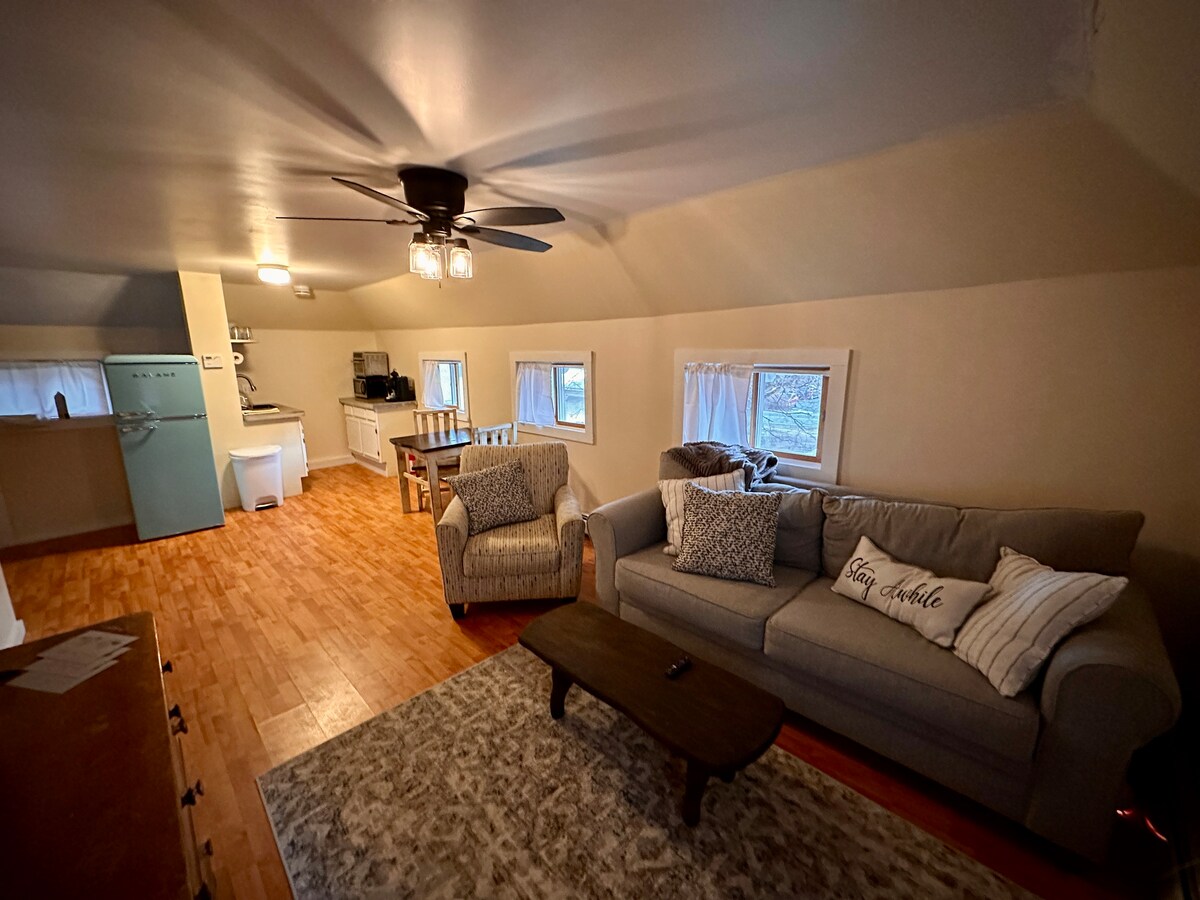 An open living space is depicted, featuring a cozy couch and an armchair with soft cushions. A wooden coffee table is situated in front, while a small dining area can be seen in the background. Natural light streams in through multiple windows, enhancing the welcoming atmosphere.
