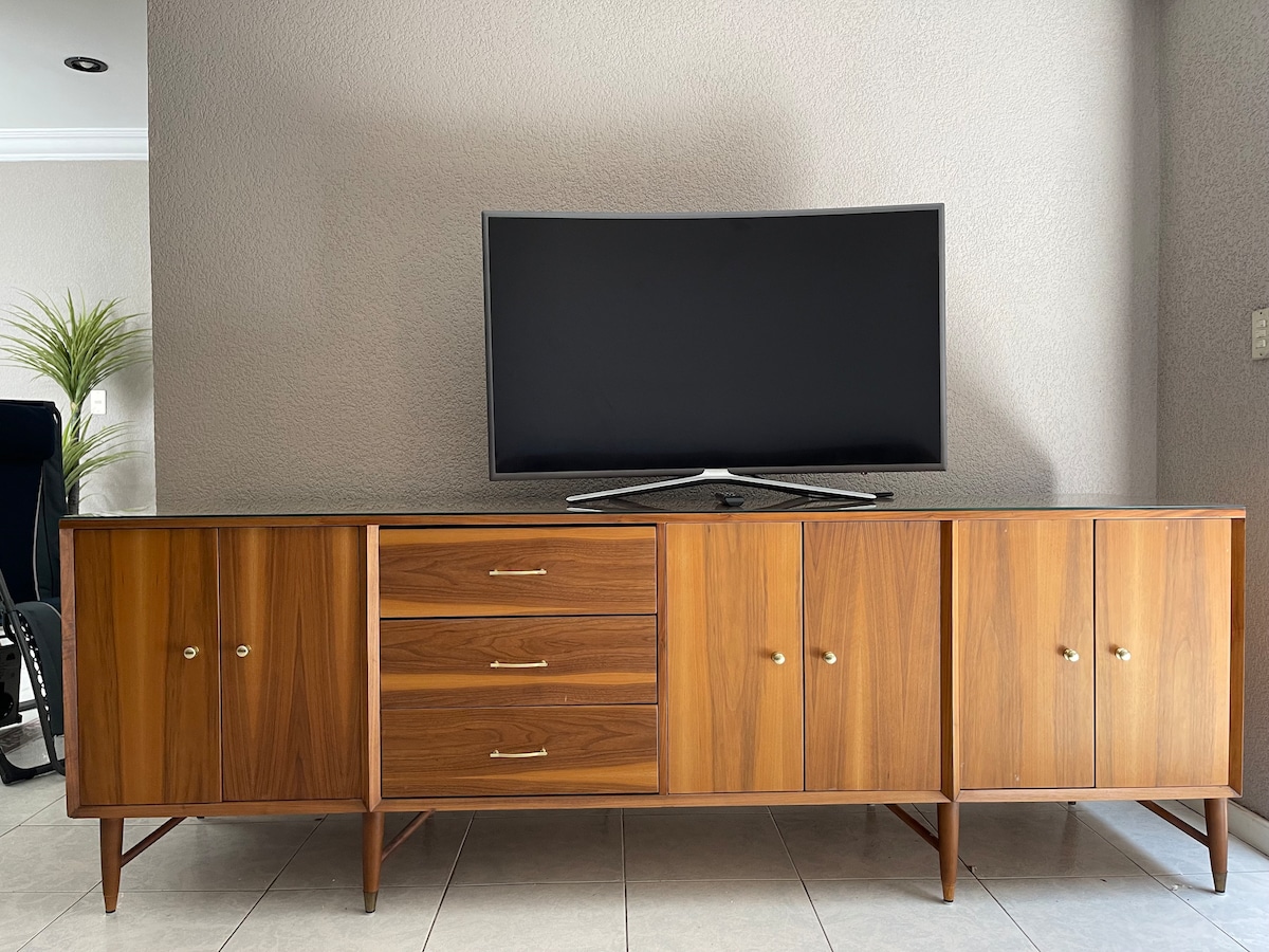 A sleek wooden cabinet with a polished finish is positioned against a neutral wall. A large flat-screen television is mounted above it. The cabinet features three drawers and several doors, providing ample storage. A decorative plant stands nearby, adding a touch of greenery.