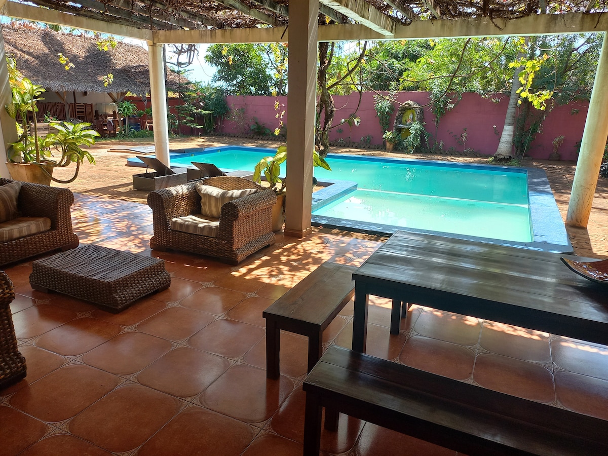 A shaded outdoor space features comfortable seating arrangements alongside a spacious pool. Sun loungers are placed near the water, surrounded by lush greenery and a vibrant pink wall in the background. Natural light filters through the overhead vines, creating a serene atmosphere.