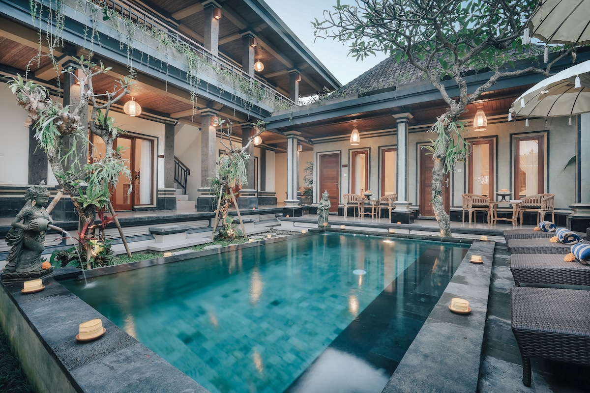 A tranquil pool area is surrounded by lush greenery and stylish seating. Two-story structures with wooden balconies frame the pool, while cozy dining sets are arranged on the patio. The scene is softly illuminated by pendant lights, creating a serene ambiance for relaxation.