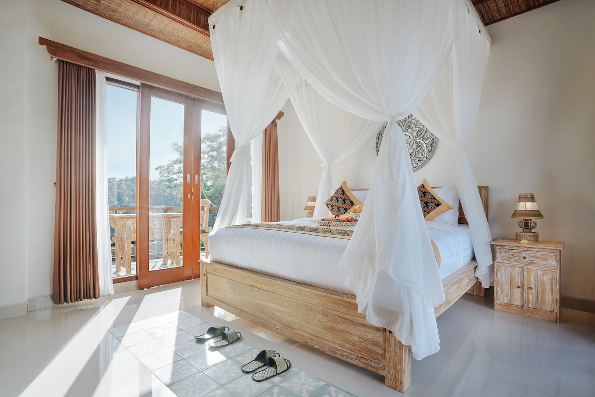 A modern double bedroom features a queen size bed draped in light curtains, complemented by two bedside tables with lamps. Expansive glass doors open to a balcony, allowing natural light to fill the space and providing views of the surrounding landscape.