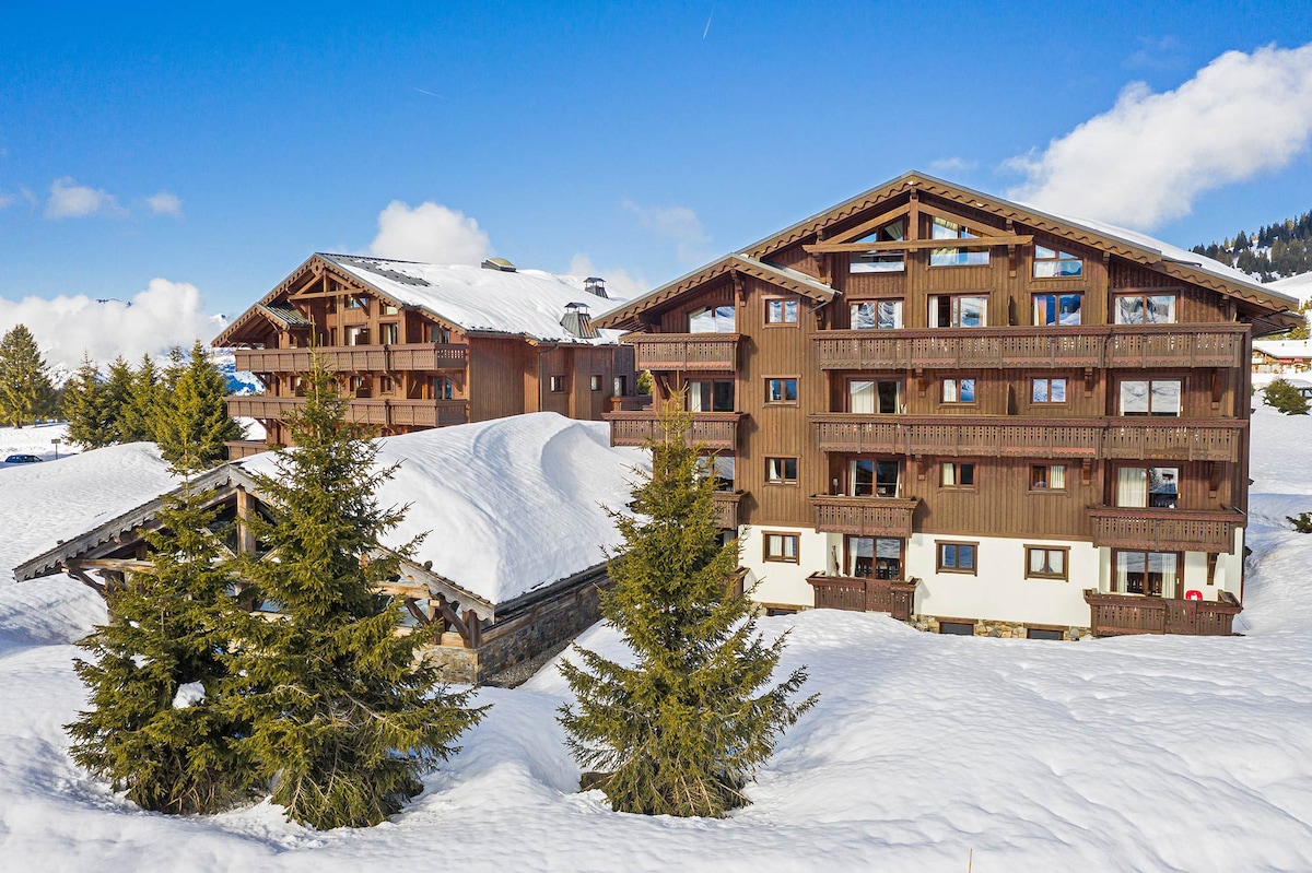 A traditional Savoyard residence is set against a snowy landscape, featuring wooden balconies and sloped roofs. The surrounding area is blanketed in snow, with evergreens visible in the foreground. A clear blue sky provides a vibrant backdrop to the charming architecture.