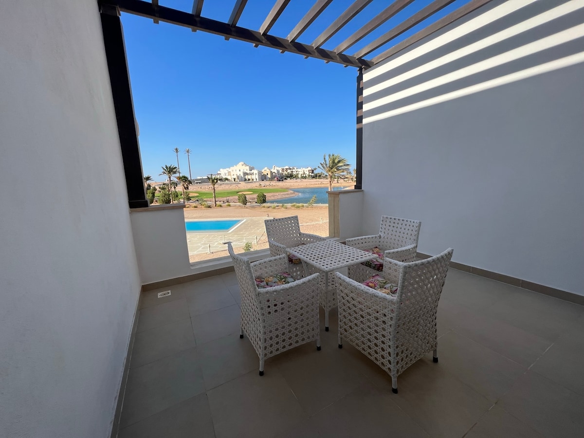 A covered terrace is furnished with a white, woven dining set, featuring a table and four chairs. Views of a pool and palm trees beyond are visible through an open wall, allowing natural light to fill the space.