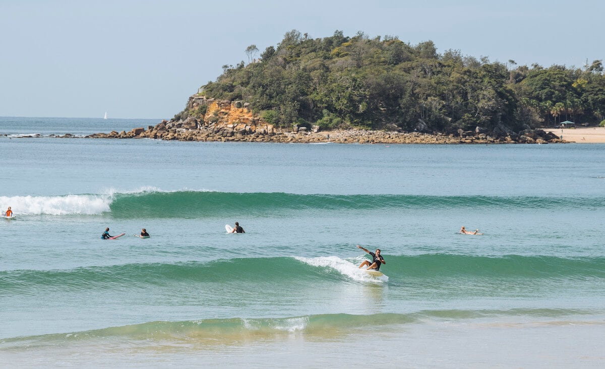 The image captures a serene beach scene with gentle waves rolling onto the shore. Surfers are seen riding the waves, while others float nearby. In the background, lush green hills rise from the water, adding to the coastal charm.