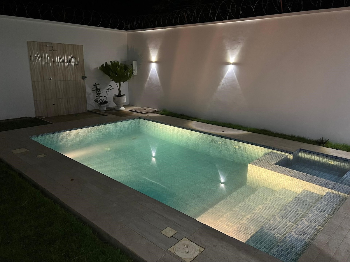 A private pool is illuminated by soft lighting against a white wall. Surrounding greenery adds to the tranquil environment. The pool area features minimalistic design elements with clear water reflecting the ambient light.