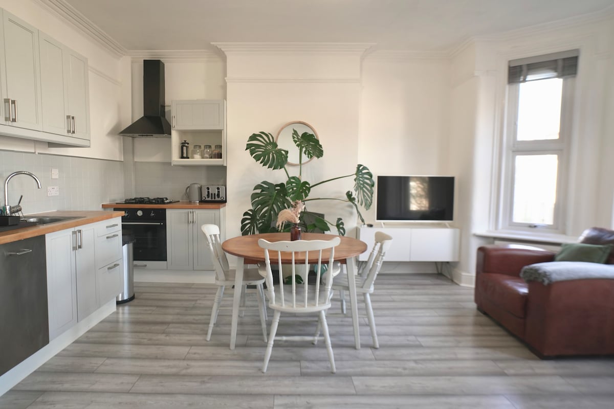 An open-plan kitchen and living area features a round wooden dining table surrounded by six white chairs. A cozy brown leather sofa is positioned nearby. Large windows illuminate the space, complemented by a green potted plant and modern appliances in the kitchen.