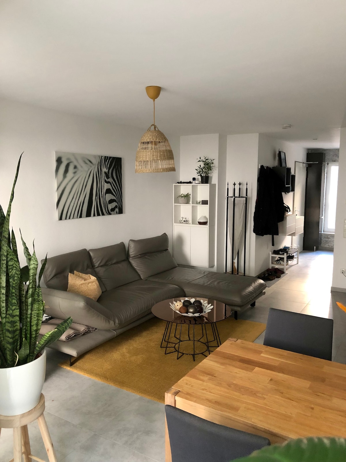 A modern living area features a sleek couch and a round coffee table set on a light area rug. A dining area with a wooden table and four chairs is visible. Green plants add a touch of nature, while wall art and a woven pendant light enhance the space.