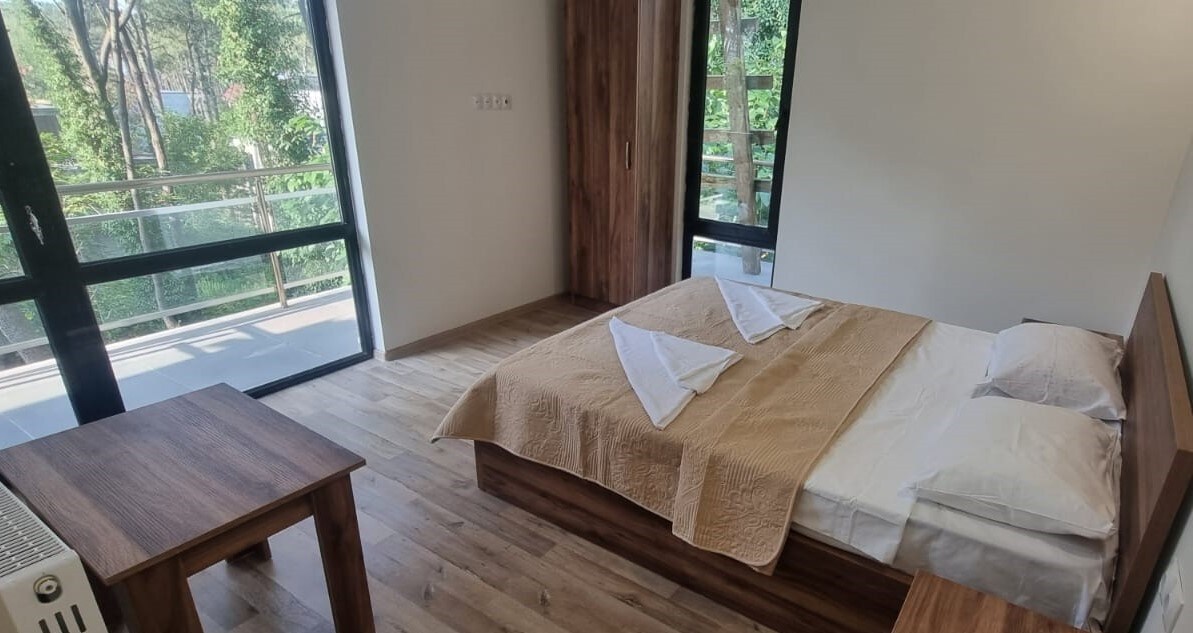 A bright sleeping area is presented with a spacious bed dressed in neutral linens. Large windows allow natural light to fill the room, revealing views of greenery. A wooden table complements the decor, enhancing a simple and comfortable atmosphere.
