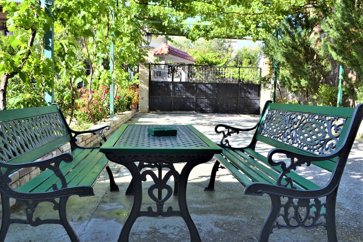 A shaded outdoor seating area features two ornate green benches positioned around a matching table. Vines and greenery create a tranquil environment, with a gated entrance visible in the background, providing a sense of privacy.