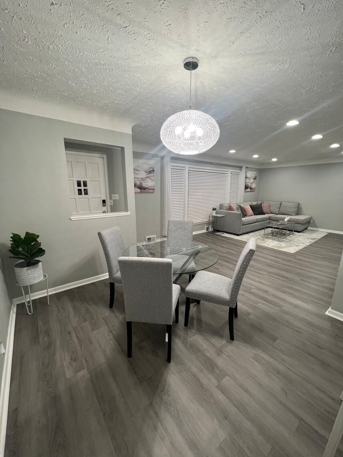 A dining area features a round glass table surrounded by four light gray upholstered chairs. The space is illuminated by a modern chandelier. A cozy seating arrangement with cushions is visible in the background, along with a decorative area rug on the floor.